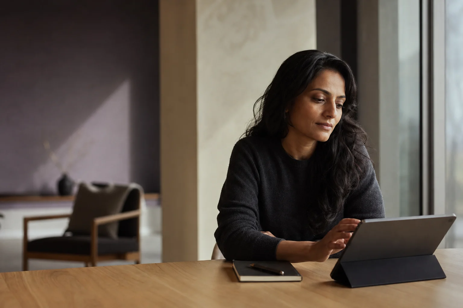 Illustrative editorial scene of an adult professional thoughtfully using a tablet beside a window